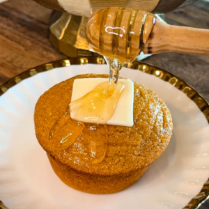 Honey being drizzled over a sweet potato honey cornbread muffin topped with a pat of butter on a decorative white plate.