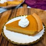 Slice of sweet potato pie with a graham cracker crust topped with whipped cream on a white plate with a whole pie blurred in the background.