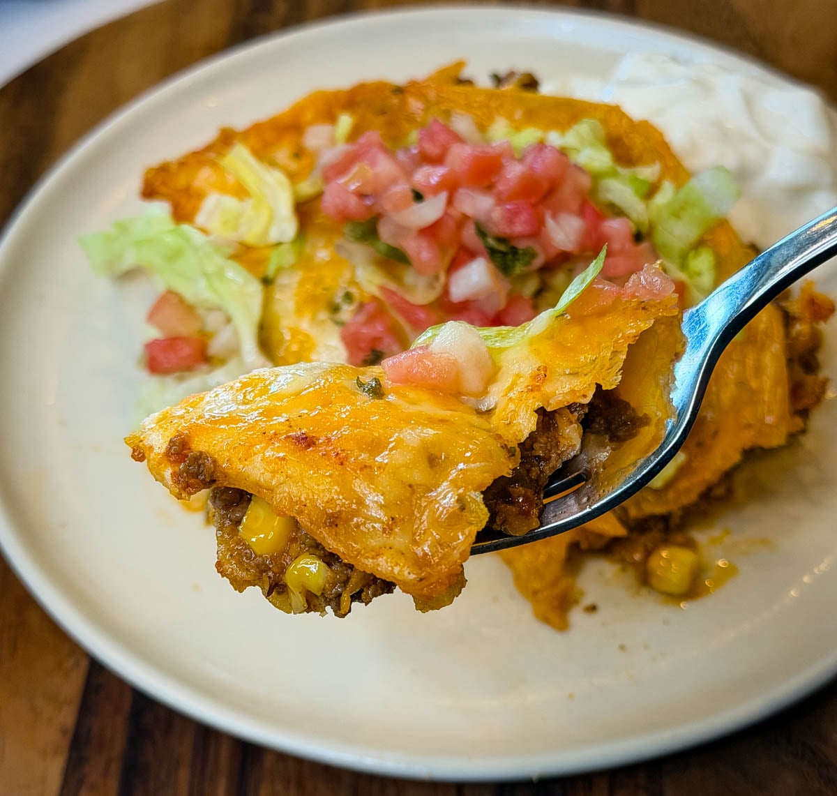 Close-up of a fork holding a bite of cheesy enchilada casserole layered with seasoned ground beef, corn, tortillas, and melted cheese, topped with pico de gallo and shredded lettuce.