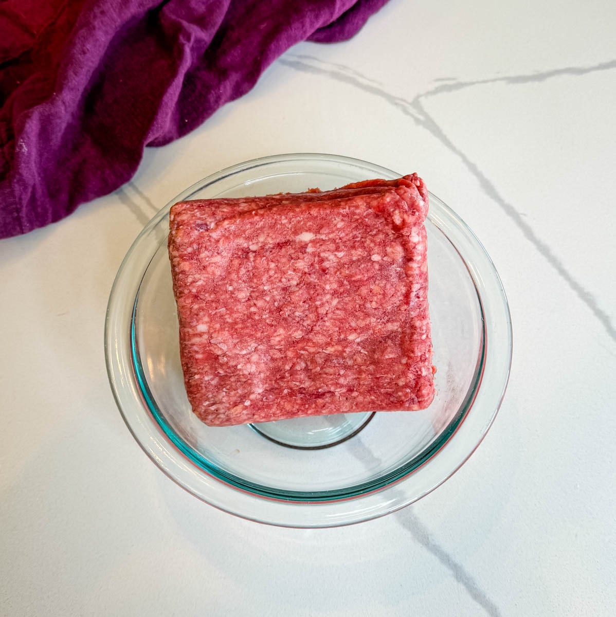 Package of raw ground beef placed in a glass bowl on a countertop, ready to be used for homemade enchilada casserole.