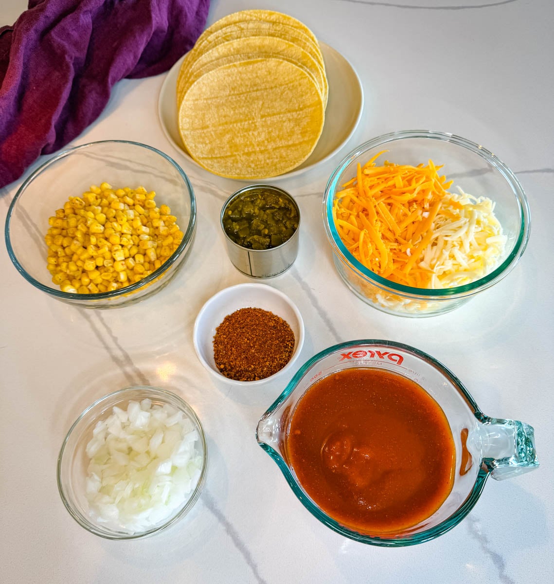 Ingredients for enchilada casserole arranged on a countertop, including ground beef, corn tortillas, enchilada sauce, shredded cheese, corn, diced onions, green chiles, and taco seasoning.