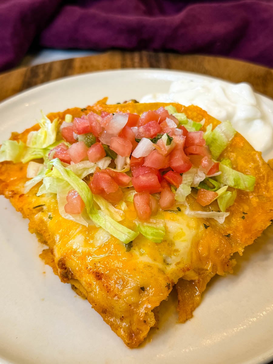 Close-up of a cheesy enchilada casserole slice topped with shredded lettuce and pico de gallo, showing the melted cheese and crispy tortilla edges.