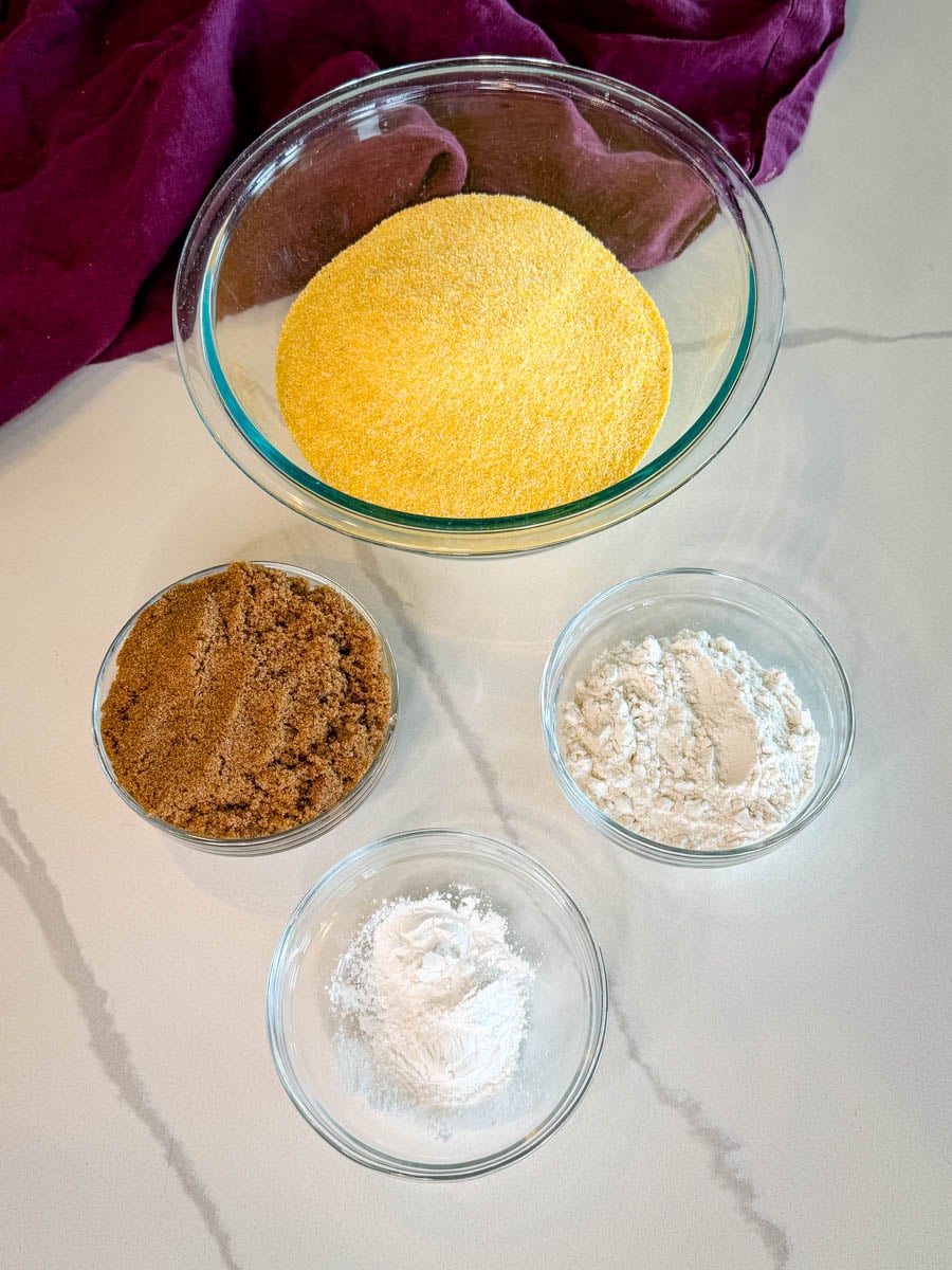 Dry ingredients for honey butter cornbread muffins including cornmeal, brown sugar, flour, and baking powder measured in glass bowls.