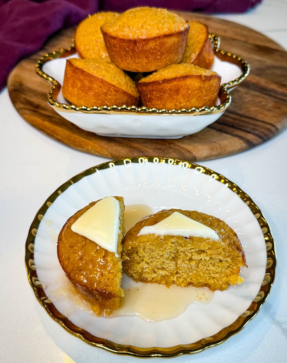 Split honey butter cornbread muffin topped with butter and honey on a decorative plate, with a dish of additional cornbread muffins in the background.