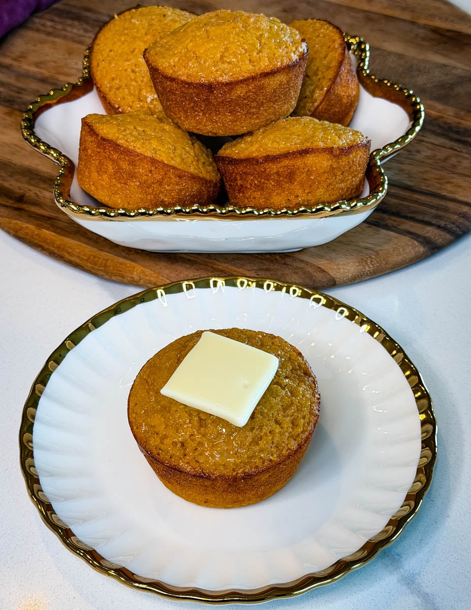 Honey butter cornbread muffin topped with butter on a white plate, with a serving dish filled with cornbread muffins in the background.