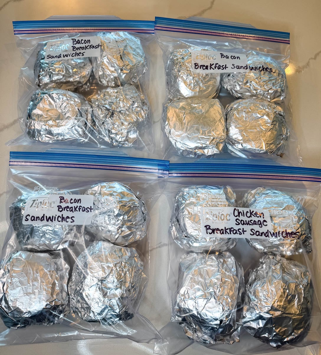 Multiple freezer breakfast sandwiches wrapped in foil and organized in labeled freezer bags with bacon and chicken sausage varieties.