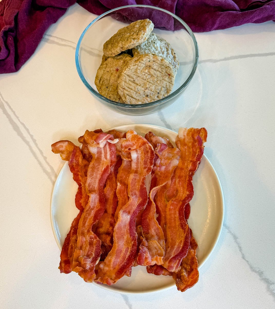 Plate of crispy cooked bacon with breakfast sausage patties in a glass bowl on a white countertop.