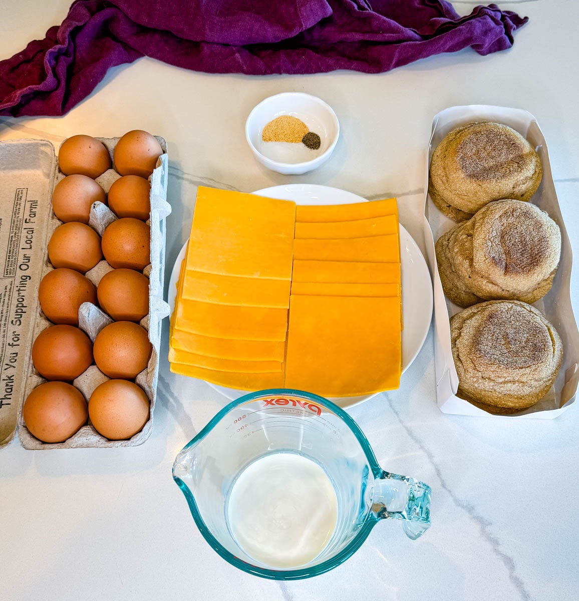 Ingredients for freezer breakfast sandwiches including eggs, sliced cheddar cheese, English muffins, milk, and a small bowl of seasonings arranged on a countertop.