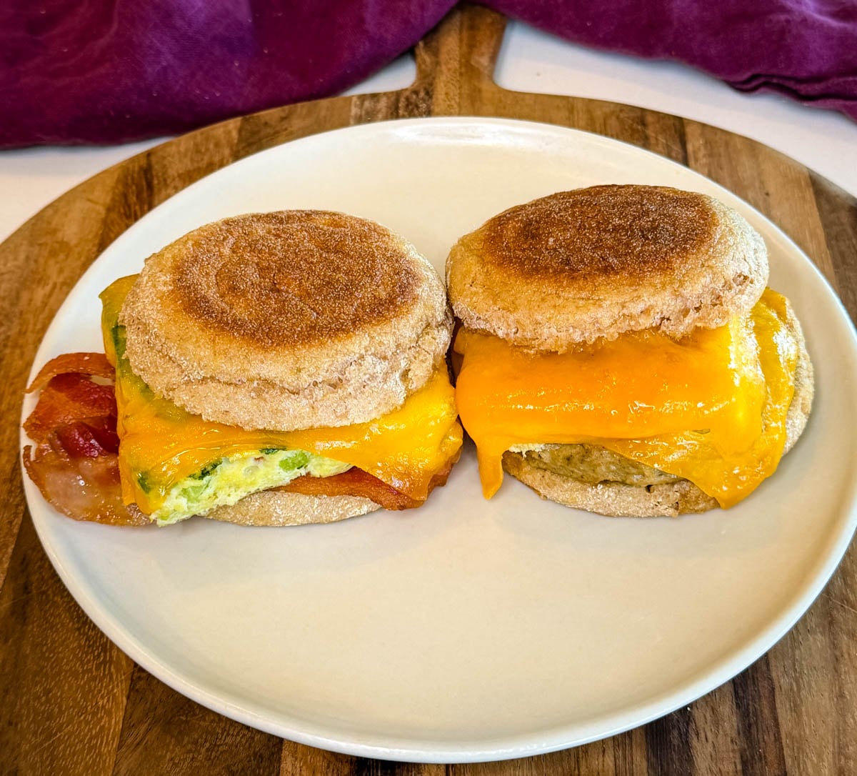 Two meal prep breakfast sandwiches with English muffins, melted cheddar cheese, eggs, bacon, and sausage served on a white plate.