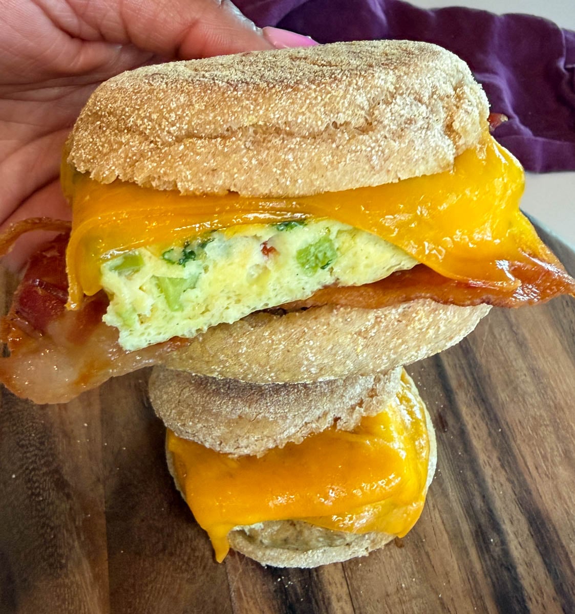 Close-up of a breakfast sandwich made with an English muffin, melted cheddar cheese, a baked egg square with vegetables, and crispy bacon.