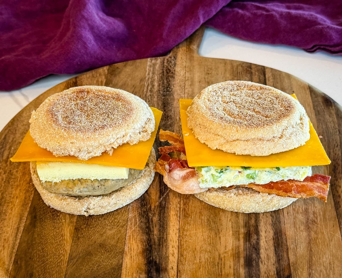 Two assembled breakfast sandwiches on a wooden serving board with English muffins, cheddar cheese, eggs, bacon, and sausage patties.