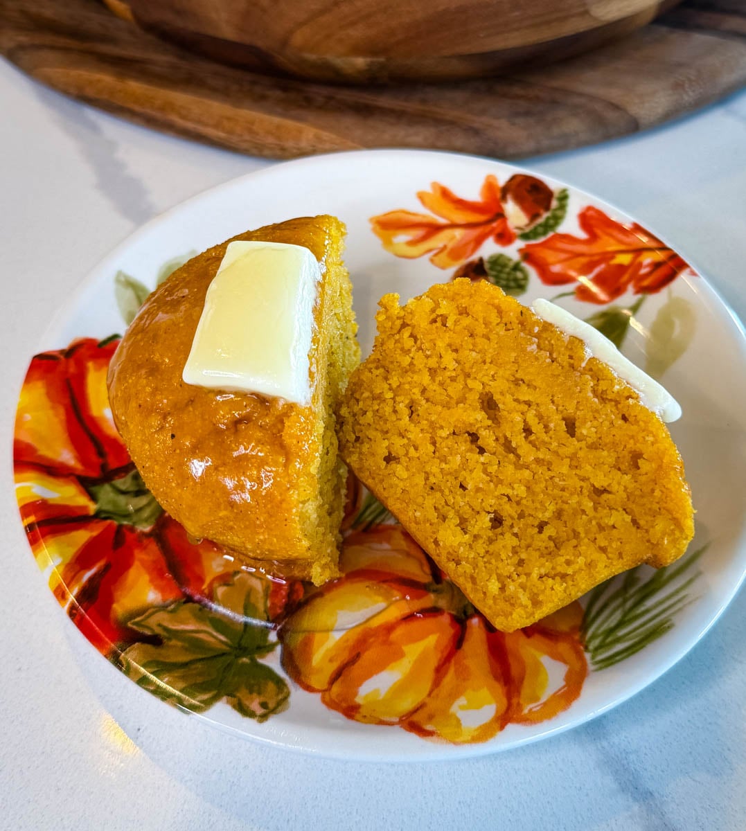 Pumpkin cornbread muffin sliced in half to show the moist, tender crumb, served with melted butter on a decorative autumn plate.