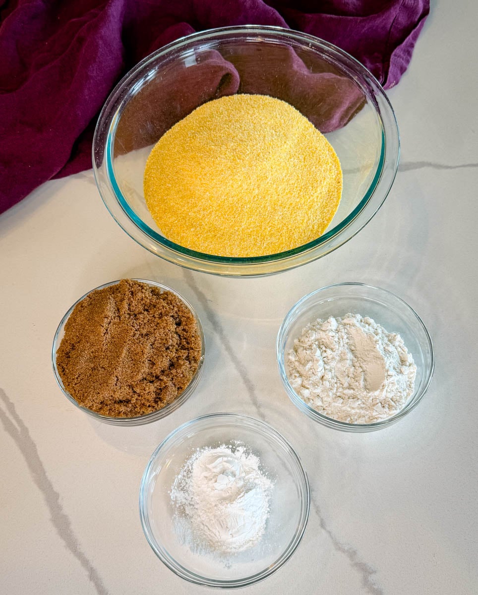 Measured dry ingredients for pumpkin cornbread muffins, including yellow cornmeal, brown sugar, flour, and baking powder arranged in glass bowls.