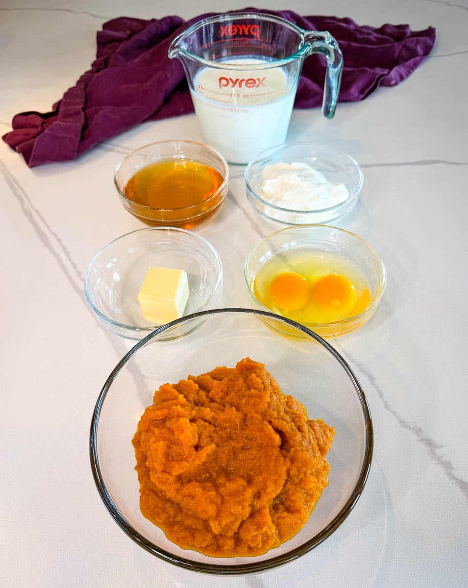 Measured wet ingredients for pumpkin cornbread muffins, including pumpkin puree, eggs, honey, butter, buttermilk, and sour cream arranged in glass bowls on a countertop.