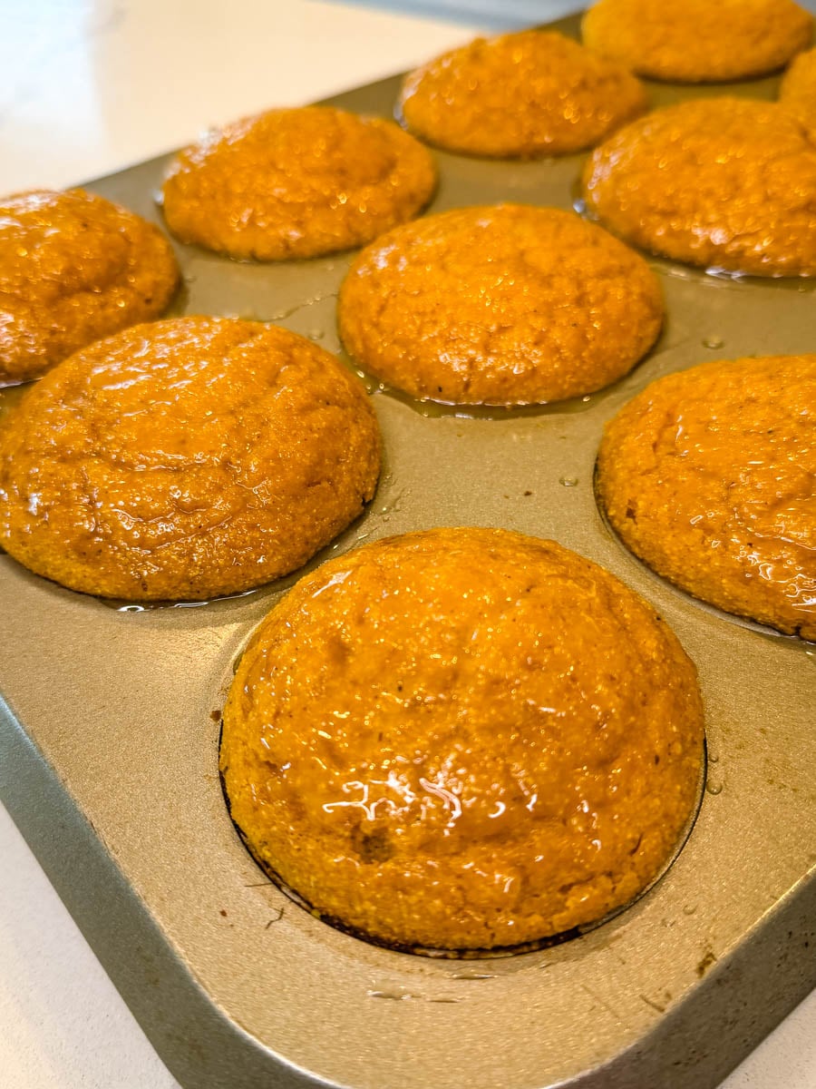 Freshly baked pumpkin cornbread muffins cooling in a muffin pan with a glossy honey glaze brushed over the tops.