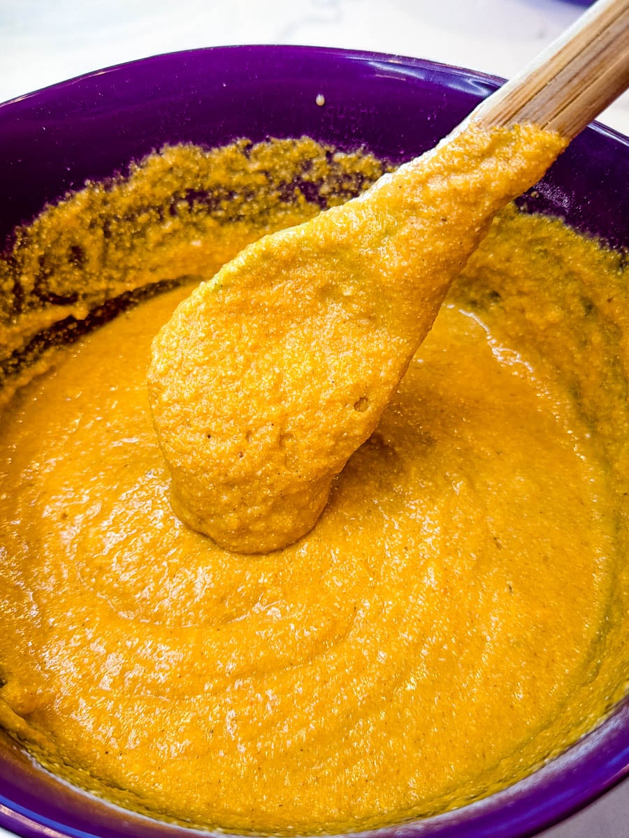 Thick pumpkin cornbread muffin batter mixed in a purple bowl, showing a smooth, rich orange texture with a spatula lifting the batter before baking.