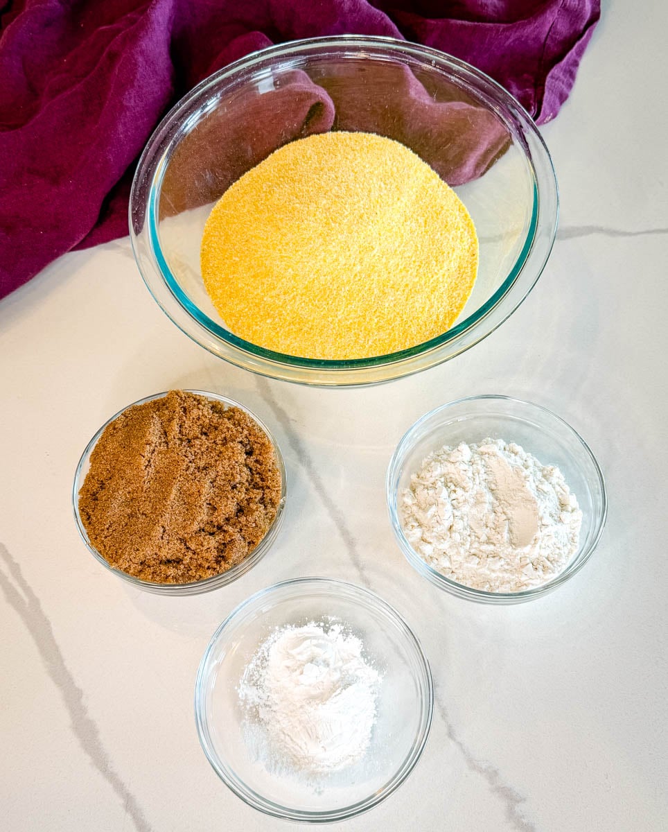 Dry ingredients for sweet potato honey cornbread muffins including yellow cornmeal, brown sugar, flour, and baking powder measured in glass bowls.