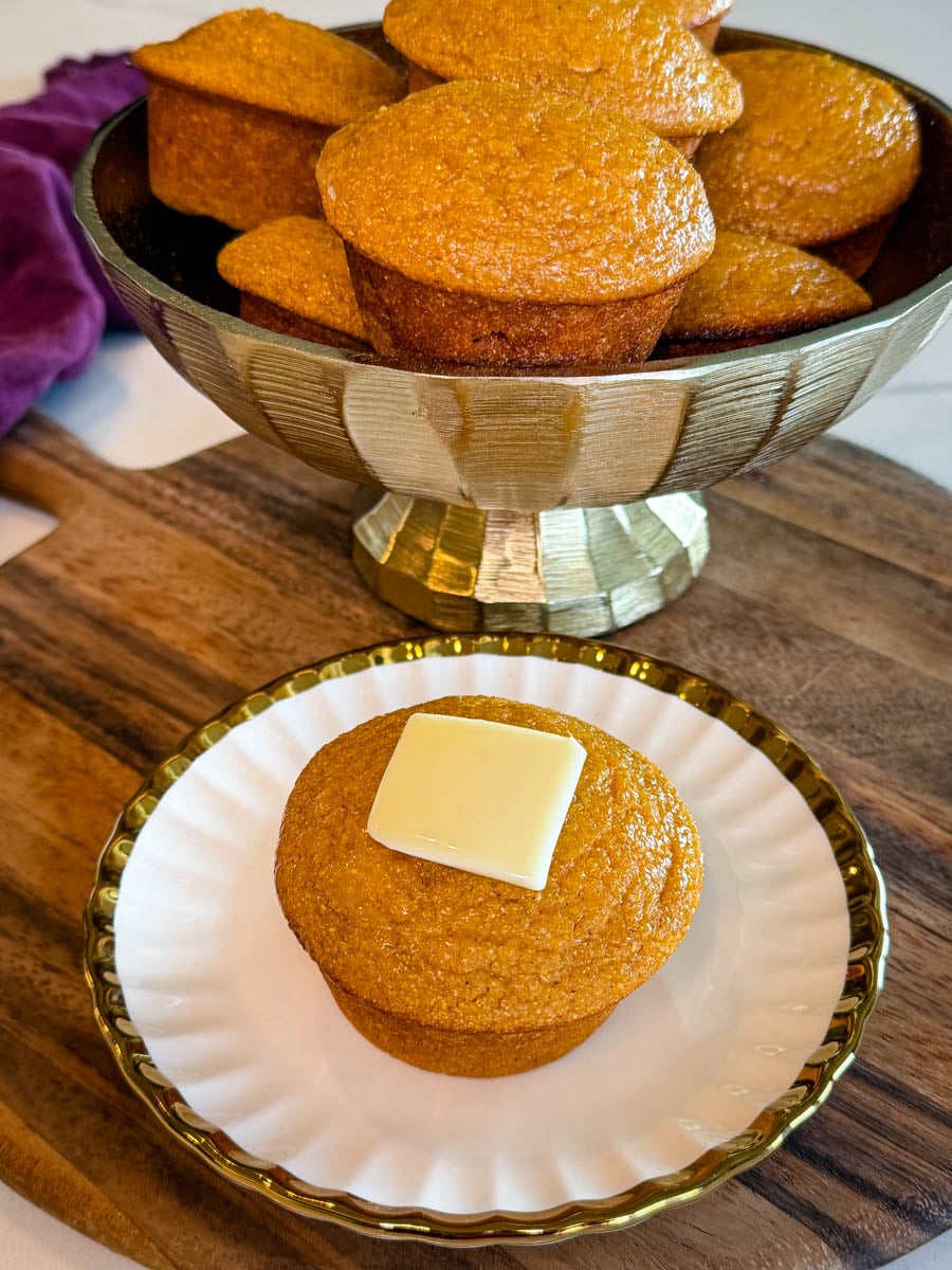 Close-up of a sweet potato honey cornbread muffin topped with butter, served in front of a bowl filled with additional muffins.