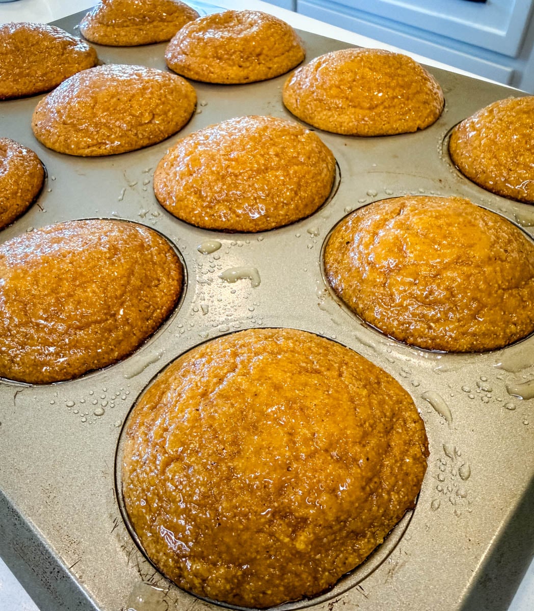Freshly baked sweet potato honey cornbread muffins in a muffin pan with a shiny honey butter glaze brushed over the tops.