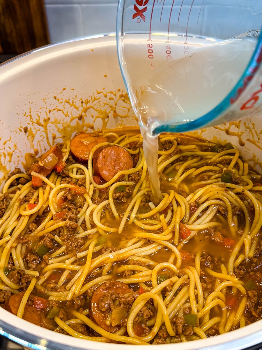 Beef broth being poured into dirty spaghetti with smoked sausage, ground beef, peppers, and onions while cooking in a pot.