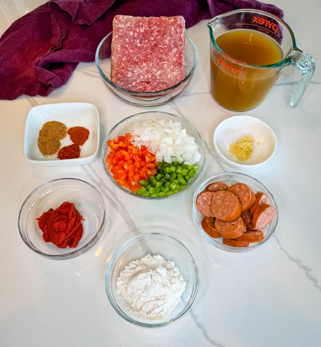 Ingredients for dirty spaghetti including ground beef, smoked sausage, beef broth, tomato paste, diced onion, bell peppers, garlic, flour, and seasonings arranged on a countertop.