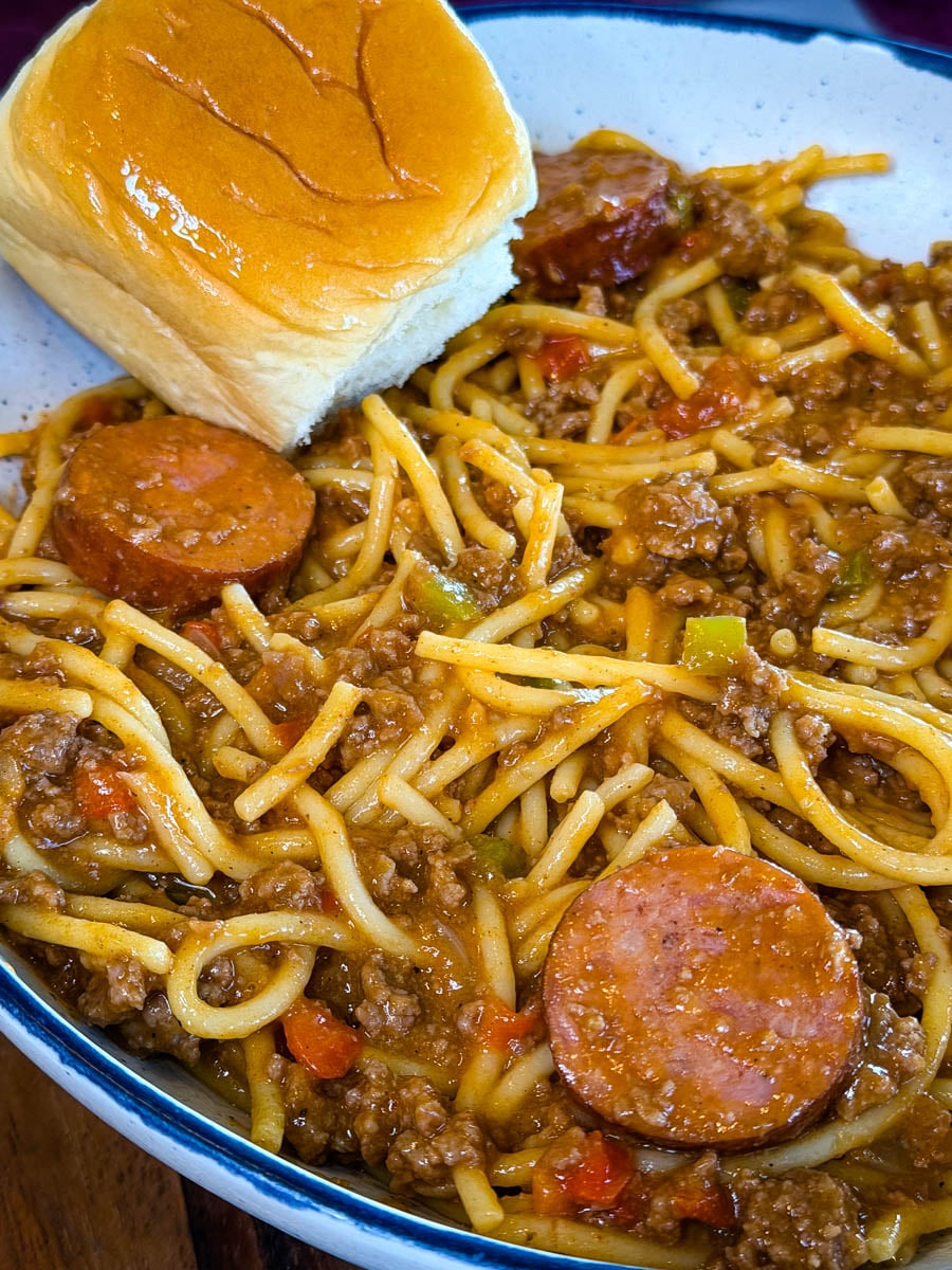 Close-up of dirty spaghetti loaded with smoked sausage, seasoned ground beef, bell peppers, onions, and a buttery Hawaiian roll.