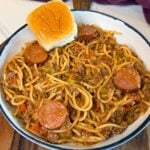 Bowl of dirty spaghetti made with ground beef, smoked sausage, bell peppers, onions, and a rich tomato-based sauce, served with a buttery Hawaiian roll.