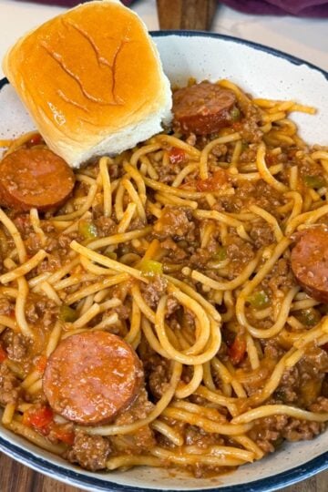 Bowl of dirty spaghetti made with ground beef, smoked sausage, bell peppers, onions, and a rich tomato-based sauce, served with a buttery Hawaiian roll.