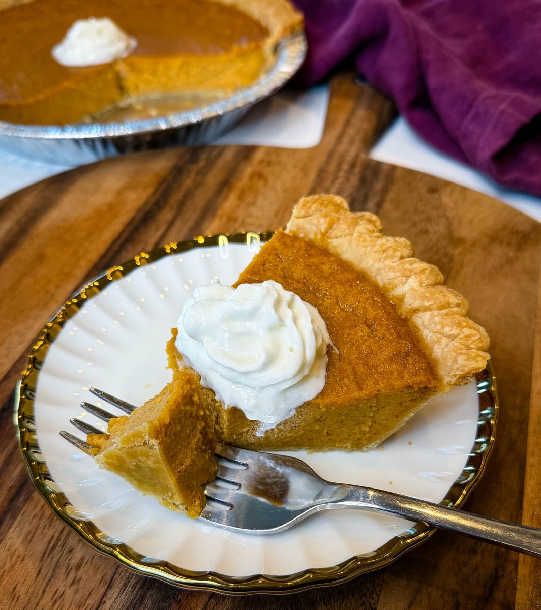 Slice of Southern sweet potato pie with a flaky store-bought crust and whipped cream, with a bite cut from the slice and the remaining pie in the background.