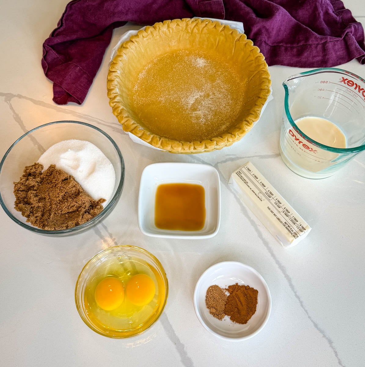 Ingredients for Southern sweet potato pie arranged on a countertop, including a frozen pie crust, sugars, evaporated milk, butter, eggs, vanilla extract, cinnamon, and nutmeg.
