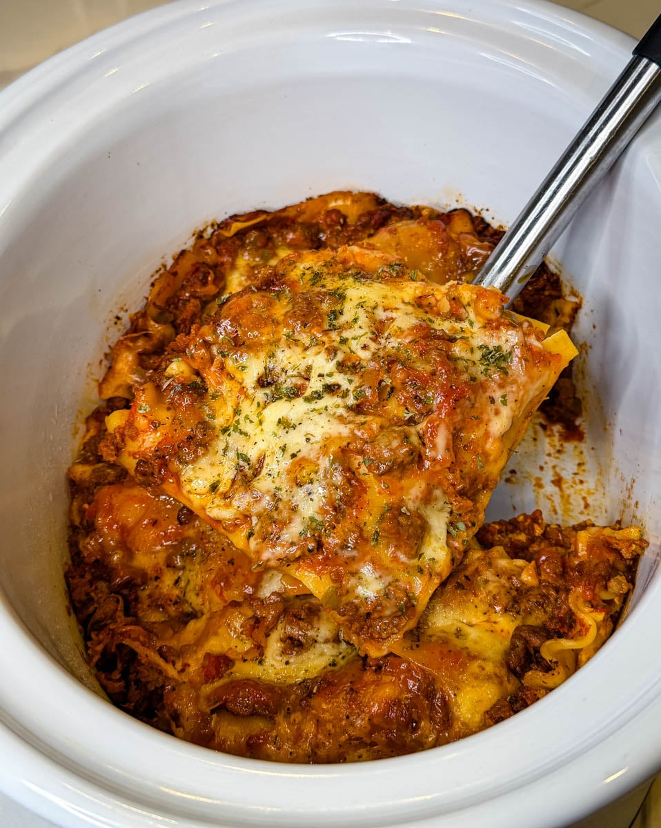 Homemade Crockpot lasagna with layers of pasta, seasoned meat sauce, and melted cheese being lifted from a white slow cooker with a serving spatula.