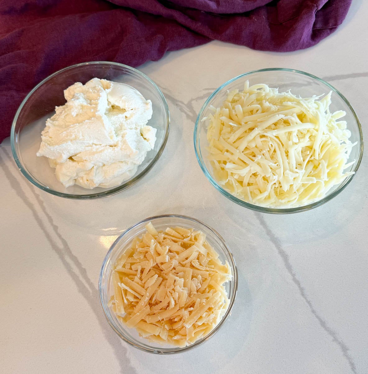 Ricotta cheese, shredded mozzarella, and shredded Parmesan cheese in separate glass bowls for making Crockpot lasagna.