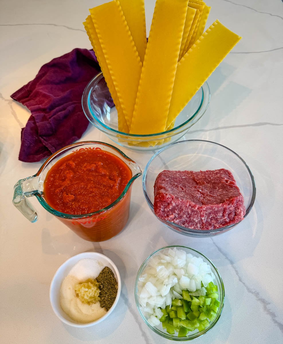 Slow cooker lasagna ingredients including uncooked lasagna noodles, ground beef, marinara sauce, diced onion, green bell pepper, garlic, and seasonings.