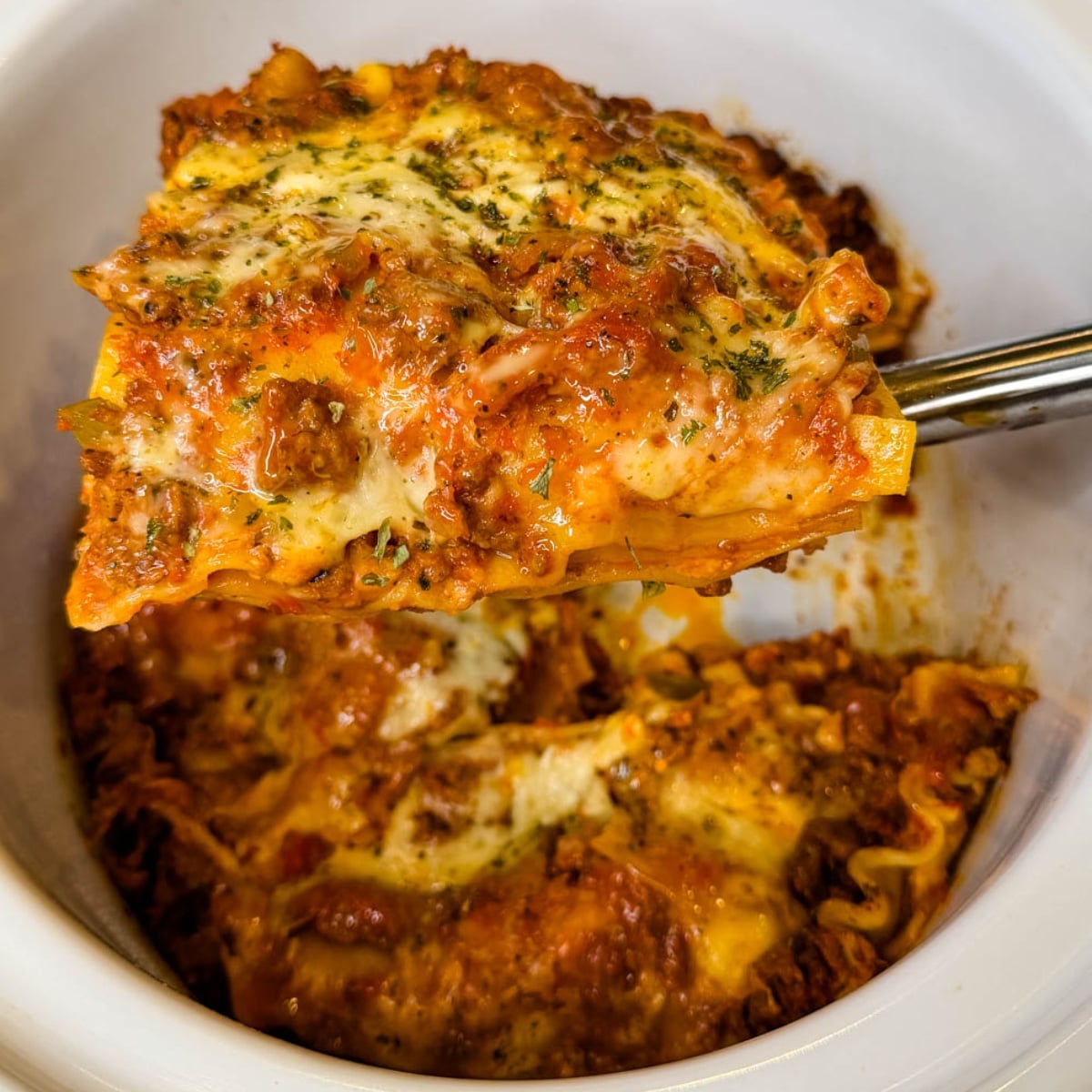 Close-up of a thick slice of slow cooker lasagna with layers of pasta, meat sauce, and melted cheese being lifted from the Crockpot.
