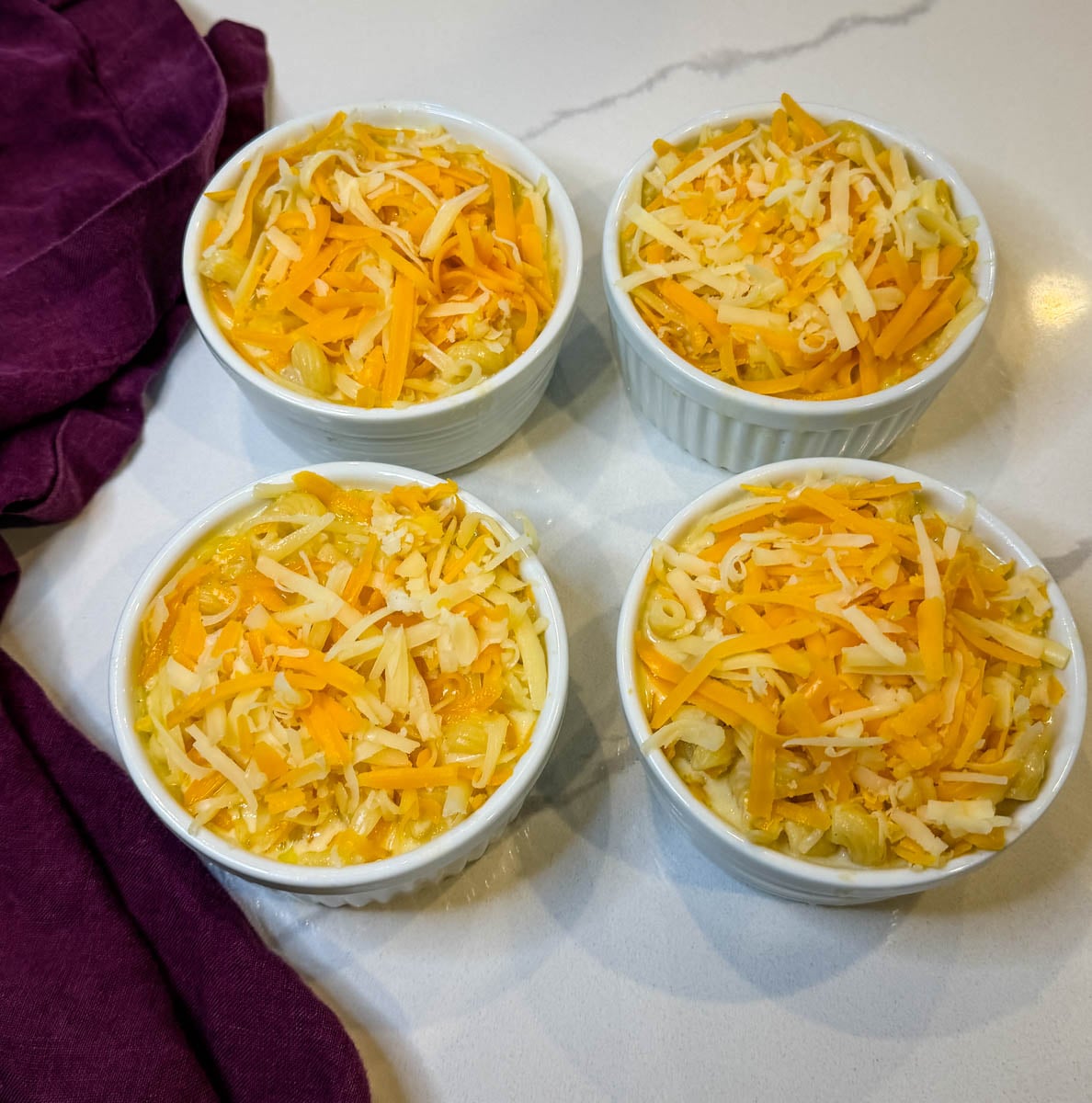 Four white ramekins filled with macaroni and cheese and a generous layer of shredded cheese, ready to cook.