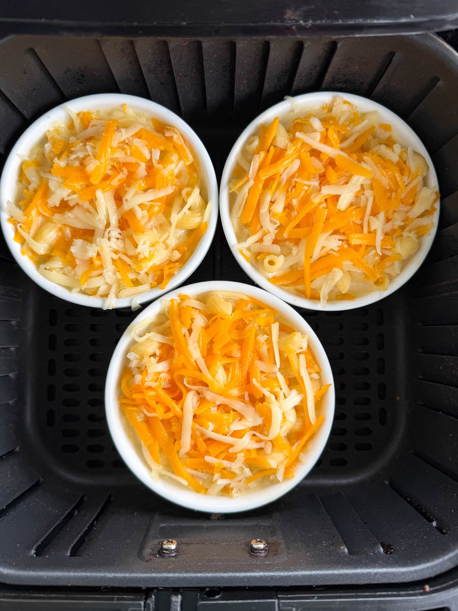 Three ramekins of macaroni and cheese topped with shredded cheddar cheese inside an air fryer basket before cooking.