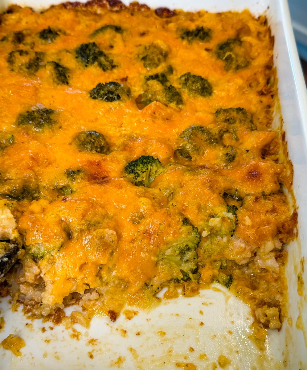 Close-up of cheesy chicken broccoli rice casserole with melted cheddar cheese and broccoli florets, with a portion scooped from the baking dish.