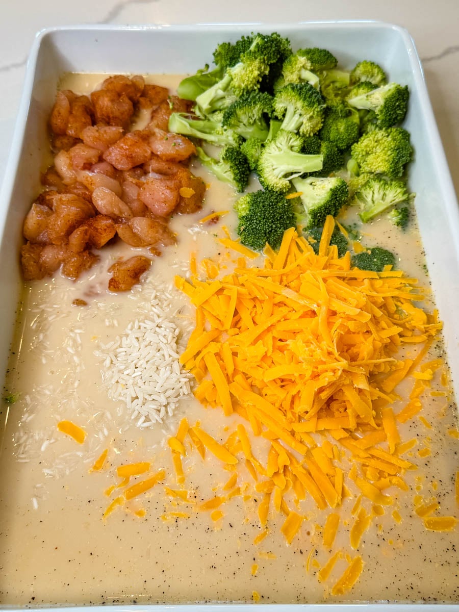 Dump-and-bake chicken broccoli rice casserole ingredients layered in a baking dish with seasoned chicken, fresh broccoli, uncooked rice, creamy sauce, and shredded cheddar cheese.