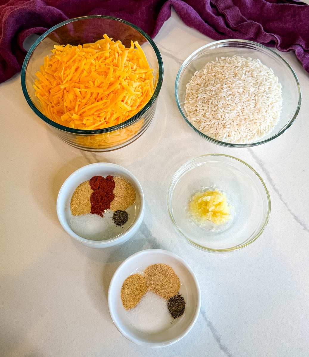 Ingredients for chicken broccoli rice casserole including shredded cheddar cheese, uncooked white rice, minced garlic, and bowls of seasonings.