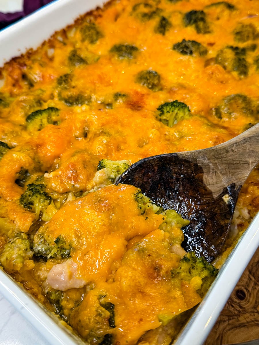 Wooden spoon serving cheesy chicken broccoli rice casserole from a white baking dish filled with chicken, broccoli, rice, and melted cheddar.