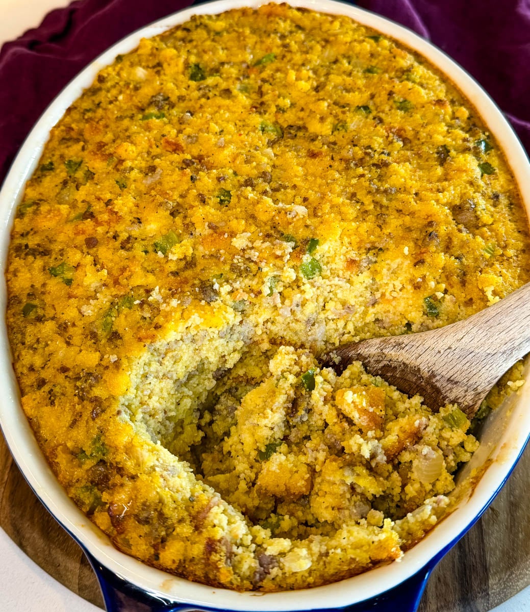 Baked cornbread dressing with sausage and vegetables in a casserole dish with a serving spoon revealing the moist interior.