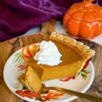 Slice of pumpkin pie with a flaky golden crust and whipped cream, served on a fall floral plate with an orange pumpkin dish in the background.