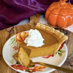 Slice of pumpkin pie with a flaky golden crust and whipped cream, served on a fall floral plate with an orange pumpkin dish in the background.
