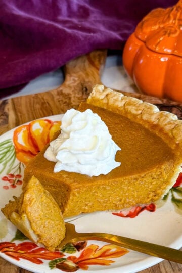 Slice of pumpkin pie with a flaky golden crust and whipped cream, served on a fall floral plate with an orange pumpkin dish in the background.