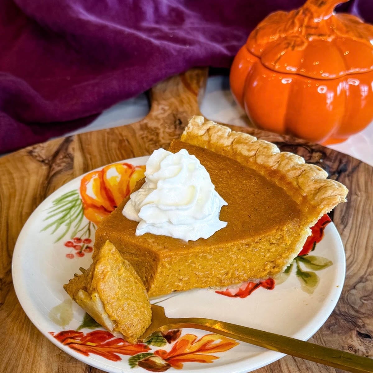 Slice of pumpkin pie with a flaky golden crust and whipped cream, served on a fall floral plate with an orange pumpkin dish in the background.