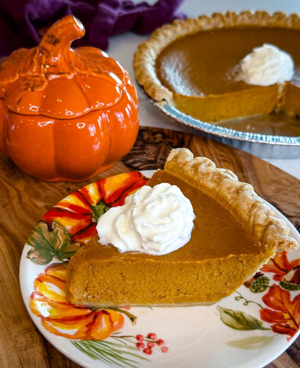 Slice of creamy pumpkin pie topped with whipped cream on a fall floral plate, with the remaining pie and an orange pumpkin dish in the background.