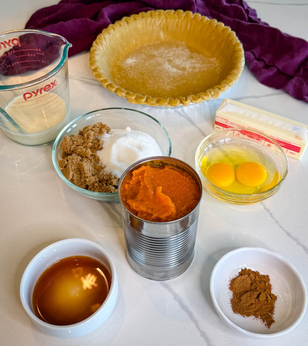 Ingredients for pumpkin pie with frozen crust, including pumpkin puree, eggs, milk, sugars, butter, spices, vanilla, and a prepared frozen pie crust.