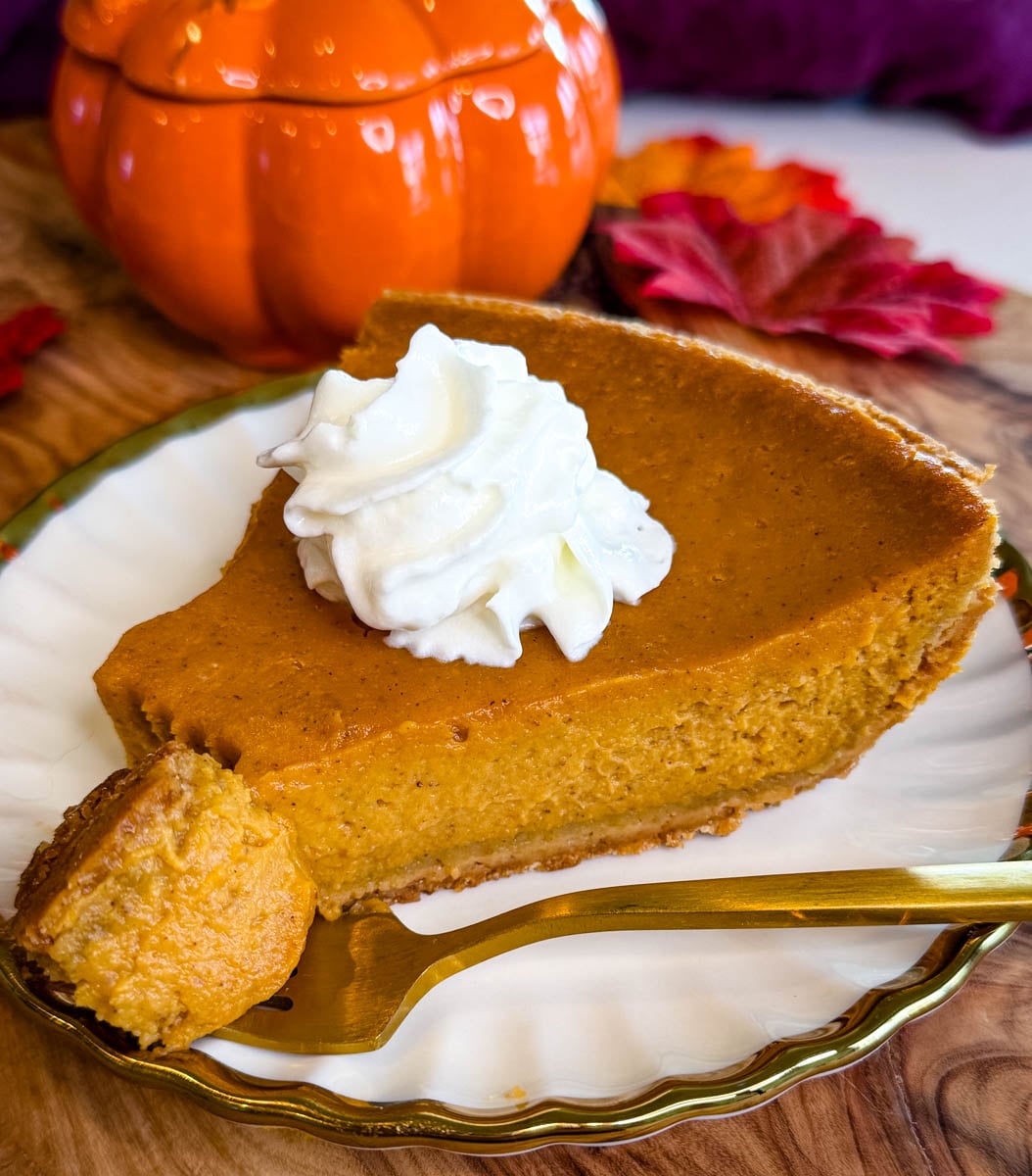 Slice of pumpkin pie with graham cracker crust topped with whipped cream and served with a gold fork.