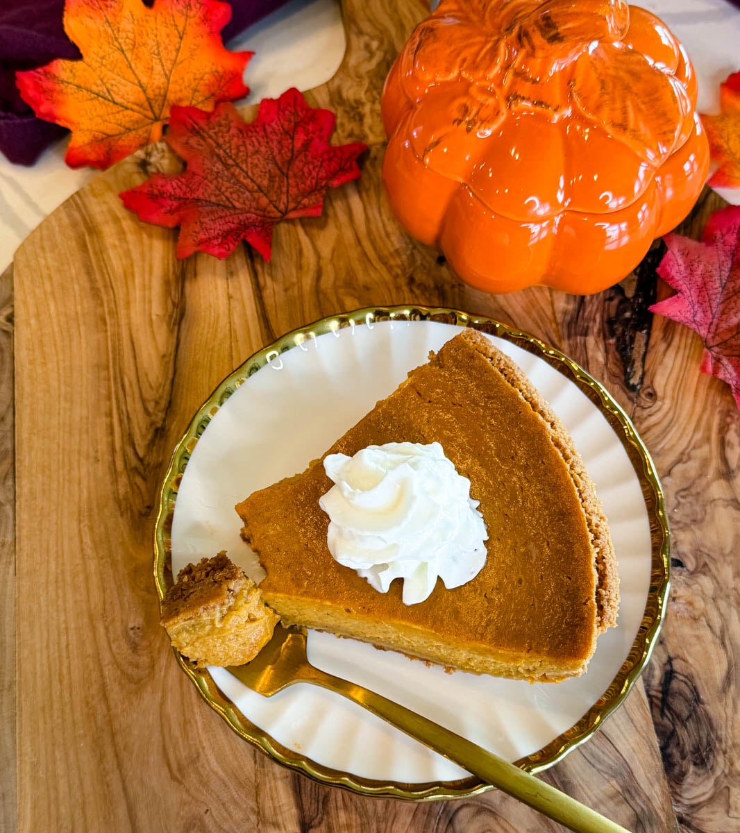 Slice of homemade pumpkin pie with graham cracker crust and whipped cream surrounded by fall leaves and pumpkin decor.