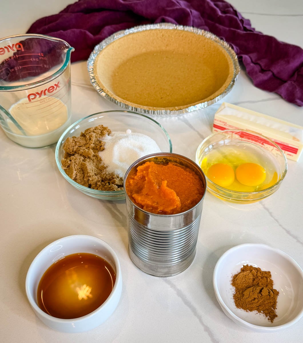 Ingredients for pumpkin pie with graham cracker crust, including pumpkin puree, eggs, milk, butter, sugars, vanilla, and warm spices.
